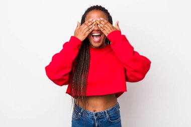 afro pretty woman smiling and feeling happy, covering eyes with both hands and waiting for unbelievable surprise