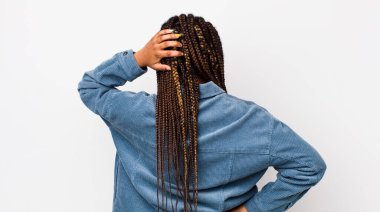 afro pretty woman feeling clueless and confused, thinking a solution, with hand on hip and other on head, rear view
