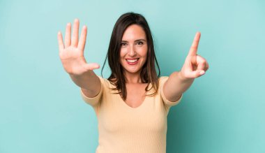young adult pretty woman smiling and looking friendly, showing number six or sixth with hand forward, counting down