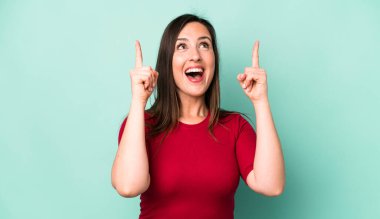 young adult pretty woman looking shocked, amazed and open mouthed, pointing upwards with both hands to copy space
