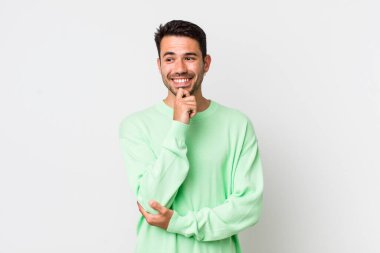 young handsome hicpanic man smiling with a happy, confident expression with hand on chin, wondering and looking to the side