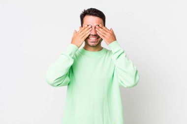 young handsome hicpanic man smiling and feeling happy, covering eyes with both hands and waiting for unbelievable surprise