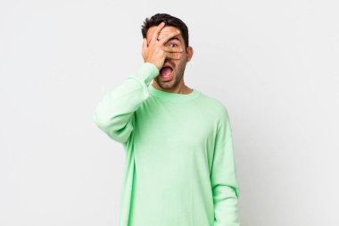 young handsome hicpanic man looking shocked, scared or terrified, covering face with hand and peeking between fingers
