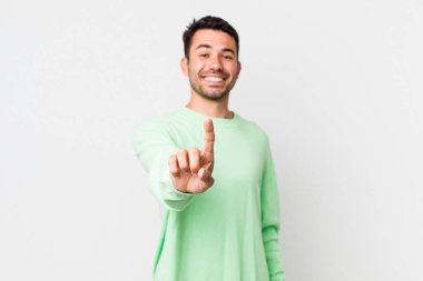 young handsome hicpanic man smiling proudly and confidently making number one pose triumphantly, feeling like a leader
