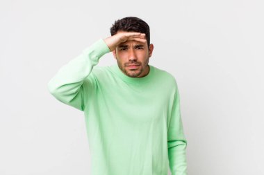 young handsome hicpanic man looking bewildered and astonished, with hand over forehead looking far away, watching or searching
