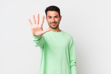 young handsome hicpanic man smiling and looking friendly, showing number five or fifth with hand forward, counting down