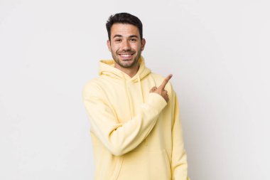 young handsome hicpanic man smiling cheerfully, feeling happy and pointing to the side and upwards, showing object in copy space