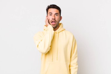 young handsome hicpanic man open-mouthed in shock and disbelief, with hand on cheek and arm crossed, feeling stupefied and amazed