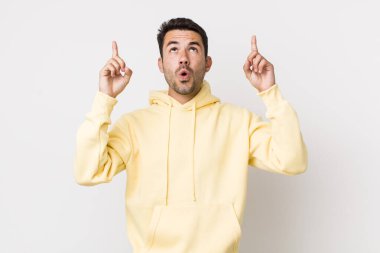 young handsome hicpanic man looking shocked, amazed and open mouthed, pointing upwards with both hands to copy space