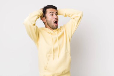 young handsome hicpanic man with open mouth, looking horrified and shocked because of a terrible mistake, raising hands to head