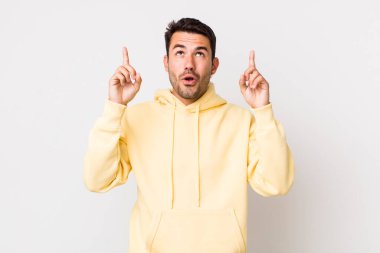 young handsome hicpanic man feeling awed and open mouthed pointing upwards with a shocked and surprised look