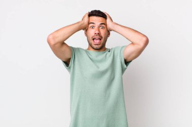 young handsome hicpanic man raising hands to head, open-mouthed, feeling extremely lucky, surprised, excited and happy