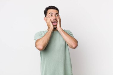 young handsome hicpanic man feeling happy, excited and surprised, looking to the side with both hands on face