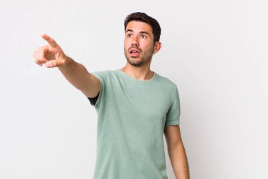 young handsome hicpanic man feeling shocked and surprised, pointing and looking upwards in awe with amazed, open-mouthed look
