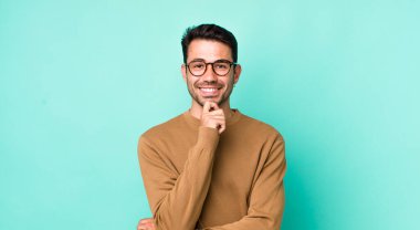 young handsome hicpanic man looking happy and smiling with hand on chin, wondering or asking a question, comparing options