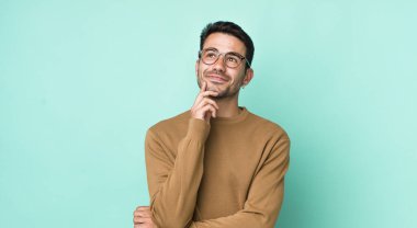 young handsome hicpanic man smiling happily and daydreaming or doubting, looking to the side