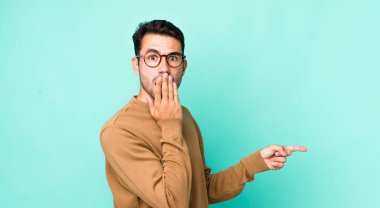 young handsome hicpanic man feeling happy, shocked and surprised, covering mouth with hand and pointing to lateral copy space