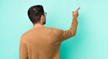 young handsome hicpanic man standing and pointing to object on copy space, rear view