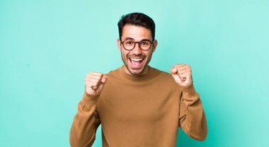 young handsome hicpanic man feeling shocked, excited and happy, laughing and celebrating success, saying wow!