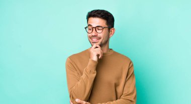 young handsome hicpanic man smiling with a happy, confident expression with hand on chin, wondering and looking to the side