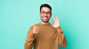 young handsome hicpanic man feeling happy, amazed, satisfied and surprised, showing okay and thumbs up gestures, smiling