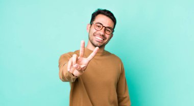 young handsome hicpanic man smiling and looking happy, carefree and positive, gesturing victory or peace with one hand