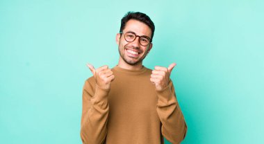 young handsome hicpanic man smiling joyfully and looking happy, feeling carefree and positive with both thumbs up