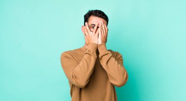young handsome hicpanic man covering face with hands, peeking between fingers with surprised expression and looking to the side
