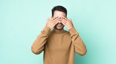 young handsome hicpanic man smiling and feeling happy, covering eyes with both hands and waiting for unbelievable surprise