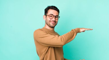 young handsome hicpanic man smiling, feeling happy, positive and satisfied, holding or showing object or concept on copy space