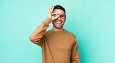 young handsome hicpanic man smiling happily with funny face, joking and looking through peephole, spying on secrets