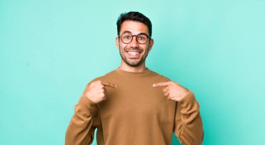 young handsome hicpanic man feeling happy, surprised and proud, pointing to self with an excited, amazed look