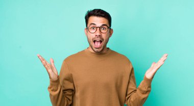 young handsome hicpanic man feeling happy, excited, surprised or shocked, smiling and astonished at something unbelievable