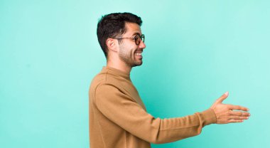young handsome hicpanic man smiling, greeting you and offering a hand shake to close a successful deal, cooperation concept