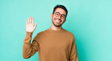 young handsome hicpanic man smiling happily and cheerfully, waving hand, welcoming and greeting you, or saying goodbye