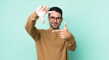 young handsome hicpanic man feeling happy, friendly and positive, smiling and making a portrait or photo frame with hands