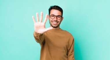 young handsome hicpanic man smiling and looking friendly, showing number five or fifth with hand forward, counting down