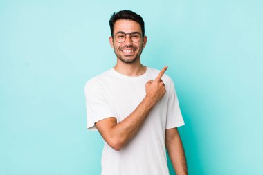 young handsome hicpanic man smiling cheerfully, feeling happy and pointing to the side and upwards, showing object in copy space