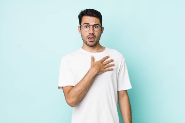 young handsome hicpanic man feeling shocked, astonished and surprised, with hand on chest and open mouth, saying who, me?