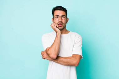 young handsome hicpanic man open-mouthed in shock and disbelief, with hand on cheek and arm crossed, feeling stupefied and amazed