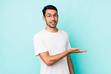 young handsome hicpanic man smiling cheerfully, feeling happy and showing a concept in copy space with palm of hand