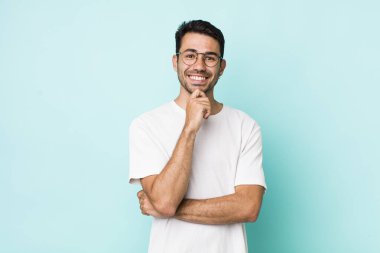 young handsome hicpanic man smiling, enjoying life, feeling happy, friendly, satisfied and carefree with hand on chin