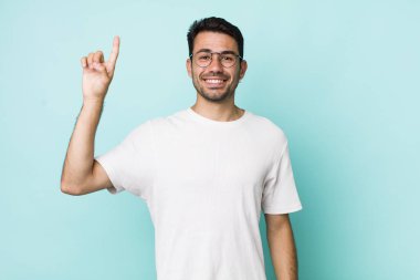 young handsome hicpanic man smiling cheerfully and happily, pointing upwards with one hand to copy space