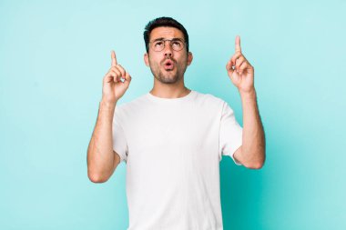 young handsome hicpanic man looking shocked, amazed and open mouthed, pointing upwards with both hands to copy space