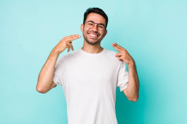 young handsome hicpanic man smiling confidently pointing to own broad smile, positive, relaxed, satisfied attitude