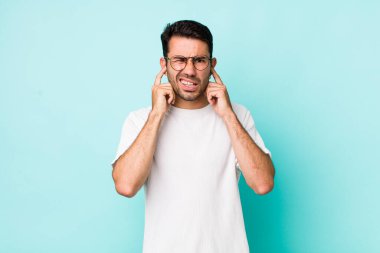 young handsome hicpanic man looking angry, stressed and annoyed, covering both ears to a deafening noise, sound or loud music