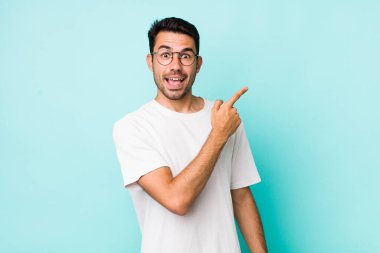 young handsome hicpanic man looking excited and surprised pointing to the side and upwards to copy space