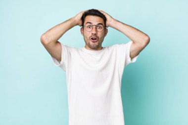 young handsome hicpanic man looking excited and surprised, open-mouthed with both hands on head, feeling like a lucky winner