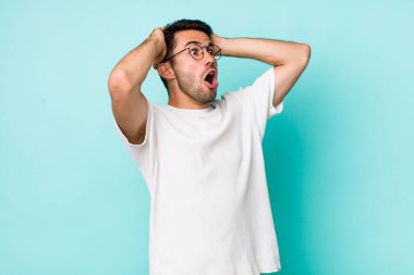 young handsome hicpanic man with open mouth, looking horrified and shocked because of a terrible mistake, raising hands to head