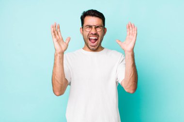 young handsome hicpanic man furiously screaming, feeling stressed and annoyed with hands up in the air saying why me